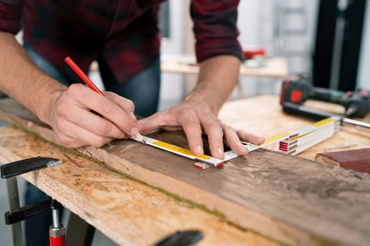 A person measuring and marking a piece of wood with a pencil and tape measure at a workstation.