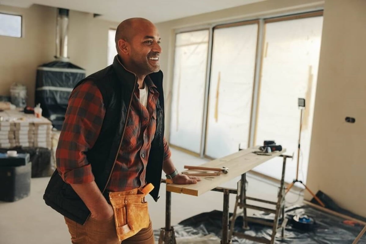 A man in a construction space wearing a tool belt and a plaid shirt, standing near renovation equipment with large windows in the background.