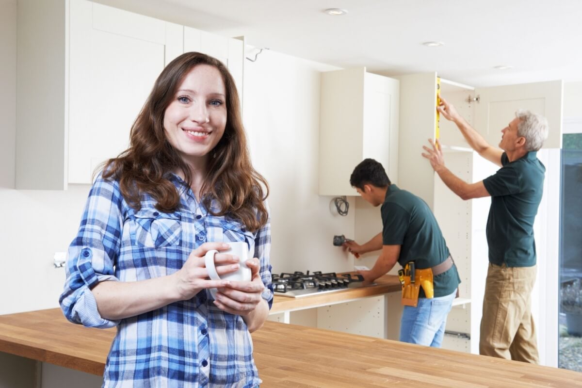 Woman in a plaid shirt holding a mug smiles at the camera while two men, using quality products, work on kitchen cabinets in the background.
