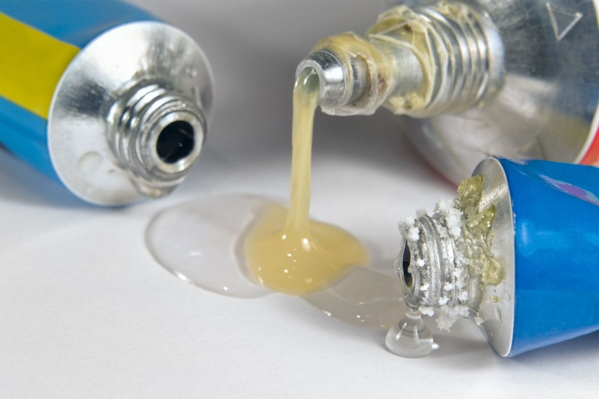 Three adhesive tubes with blue bodies, two featuring open threaded tips. Clear and yellowish substances are leaking from the tubes onto a surface.
