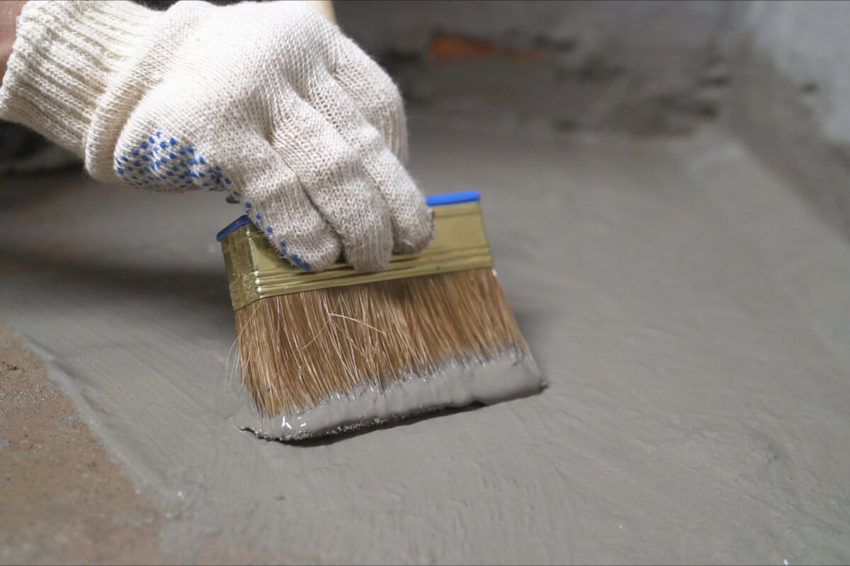 A person wearing gloves applies a gray waterproofing substance on the floor with a large brush, using quality products to ensure durability and effective protection.