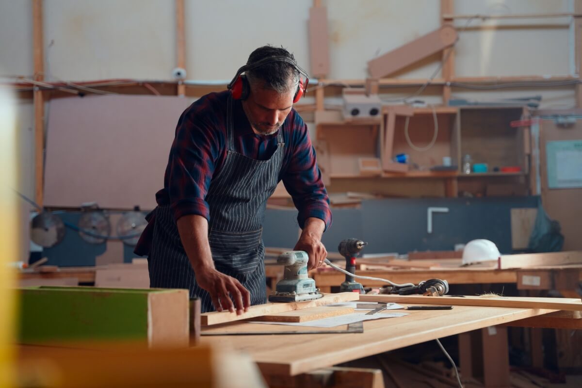 In a cluttered workshop, a man with ear protection and an apron meticulously sands a wooden plank on the workbench.